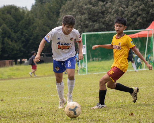 Two young soccer players in action on a grassy field, focused on controlling the ball. One wears a white and blue uniform while the other wears a yellow and red uniform. In the background, a soccer goalpost and trees are visible.