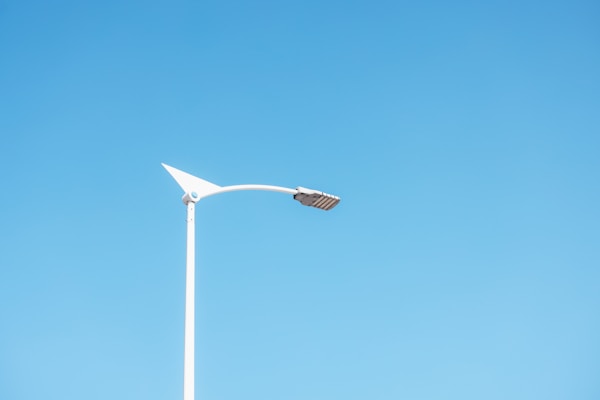 A modern streetlamp with a sleek, minimalist design is set against a clear blue sky. The lamp features an angular, triangular top and a long, slender pole.