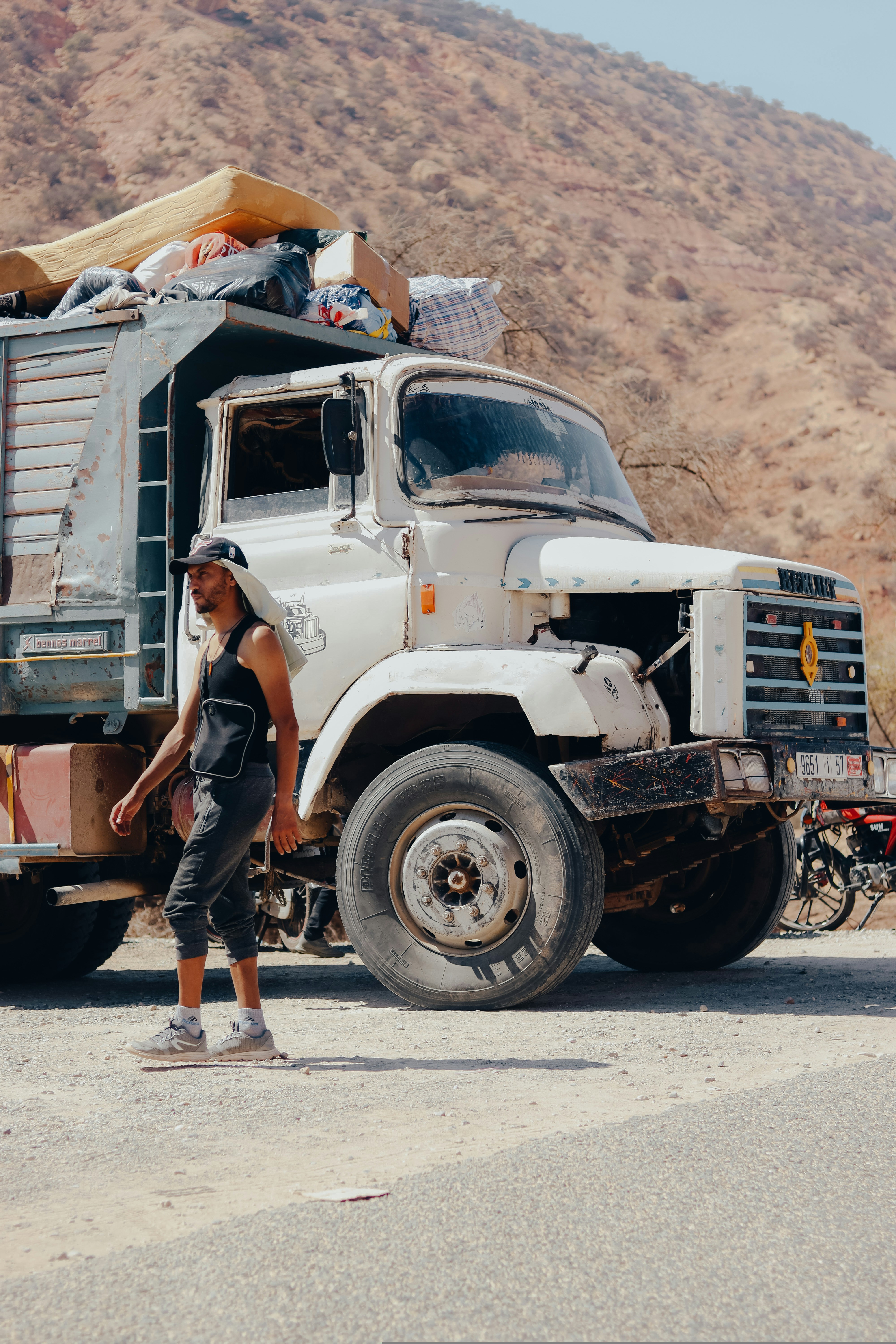 A man in casual clothing and a cap walks past an old, damaged white truck loaded with belongings, parked roadside with dry, rocky hills in the background—a stark contrast to modern tow truck leasing options.
