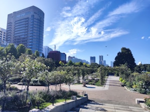 A well-maintained park featuring a variety of green shrubbery and flowering plants. The park includes paved walkways that are lined with well-trimmed bushes and small trees. In the background, there are tall modern buildings, including a hotel, set against a bright blue sky with scattered white clouds. Birds can be seen flying near the buildings. 