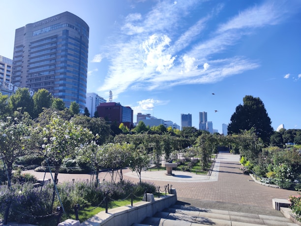 A well-maintained park featuring a variety of green shrubbery and flowering plants. The park includes paved walkways that are lined with well-trimmed bushes and small trees. In the background, there are tall modern buildings, including a hotel, set against a bright blue sky with scattered white clouds. Birds can be seen flying near the buildings. 