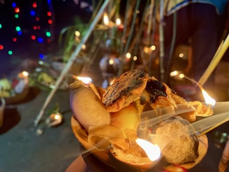 An arrangement of lit diyas with burning wicks and oil is placed in a decorative holder made of natural materials. In the background, colorful lights create a festive ambiance, while partial views of additional decorations and people suggest a cultural or religious event.
