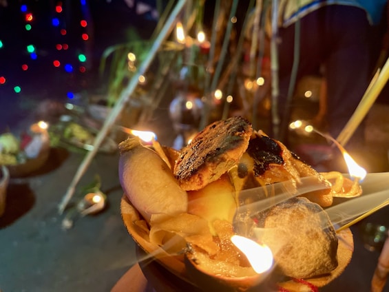 An arrangement of lit diyas with burning wicks and oil is placed in a decorative holder made of natural materials. In the background, colorful lights create a festive ambiance, while partial views of additional decorations and people suggest a cultural or religious event.