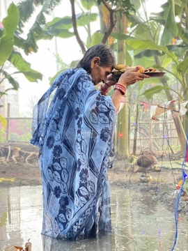 An elderly person wearing a patterned blue saree holds a tray of offerings while standing in shallow water. Surrounding the individual are green plants and decorations hanging on a string. The environment suggests a ritual or cultural ceremony taking place outdoors.
