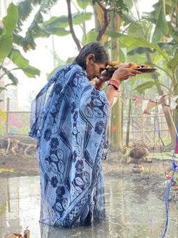 An elderly person wearing a patterned blue saree holds a tray of offerings while standing in shallow water. Surrounding the individual are green plants and decorations hanging on a string. The environment suggests a ritual or cultural ceremony taking place outdoors.
