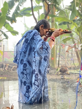 An elderly person wearing a patterned blue saree holds a tray of offerings while standing in shallow water. Surrounding the individual are green plants and decorations hanging on a string. The environment suggests a ritual or cultural ceremony taking place outdoors.