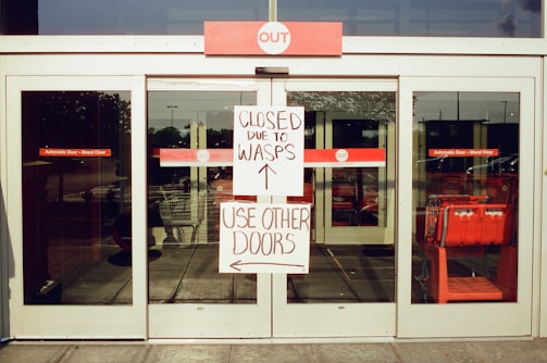 A sliding glass door with signs indicating it is closed due to wasps. The signs advise using other doors. Shopping carts are visible through the glass.