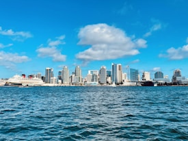 A clear blue sky with fluffy clouds frames a vibrant city skyline along the waterfront. High-rise buildings, modern and varied in design, line the horizon. In the foreground, a large cruise ship is docked near the shore, while a sailboat gently glides across the sparkling water.