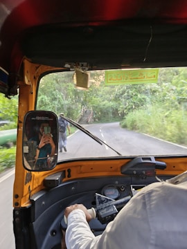 The interior of an auto rickshaw is shown with a view of a winding road ahead. The driver's arm is partially visible as they steer the vehicle. A mirror on the rickshaw displays a reflection of a passenger taking a photo with a smartphone. Lush green foliage lines the road, and a person is seen walking on the road.