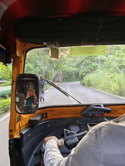 The interior of an auto rickshaw is shown with a view of a winding road ahead. The driver's arm is partially visible as they steer the vehicle. A mirror on the rickshaw displays a reflection of a passenger taking a photo with a smartphone. Lush green foliage lines the road, and a person is seen walking on the road.