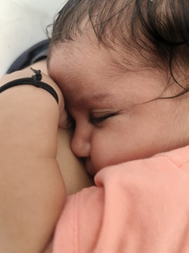 A close-up of a sleeping baby with eyes closed, resting on a soft surface. The baby is wearing a pink outfit and has a black elastic band on the wrist. The peaceful sleep and gentle positioning create a serene atmosphere.