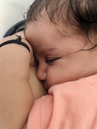 A close-up of a sleeping baby with eyes closed, resting on a soft surface. The baby is wearing a pink outfit and has a black elastic band on the wrist. The peaceful sleep and gentle positioning create a serene atmosphere.