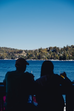 Silhouettes of two people sitting on a balcony or terrace, overlooking a large, serene lake with a tree-covered shoreline in the background. The sky is clear and blue, and one person is holding a drink.