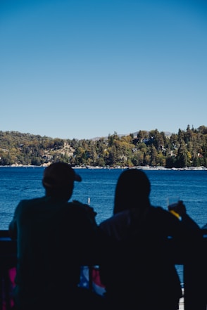 Silhouettes of two people sitting on a balcony or terrace, overlooking a large, serene lake with a tree-covered shoreline in the background. The sky is clear and blue, and one person is holding a drink.