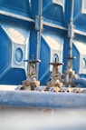 Two metallic valves with wheel handles are attached to horizontal pipes, set against a backdrop of a large blue industrial container. The setting suggests an industrial or mechanical environment with a focus on infrastructure or utilities.
