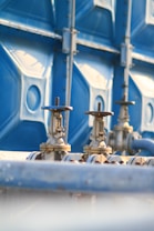Two metallic valves with wheel handles are attached to horizontal pipes, set against a backdrop of a large blue industrial container. The setting suggests an industrial or mechanical environment with a focus on infrastructure or utilities.