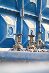 Two metallic valves with wheel handles are attached to horizontal pipes, set against a backdrop of a large blue industrial container. The setting suggests an industrial or mechanical environment with a focus on infrastructure or utilities.