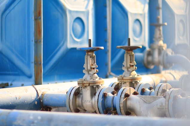 A detailed close-up of industrial pipelines and valves, with a focus on metal pipes and turning mechanisms set against a blue panel background. The metal appears slightly weathered, indicative of regular use.