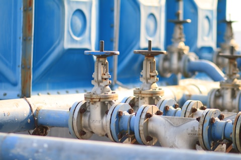 A series of industrial pipes and valves made of metal, positioned against a backdrop of large blue containers. The metal surfaces have signs of rust and wear, indicating regular use. The lighting is natural, suggesting the setting is outdoors in daylight.