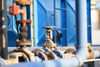 A close-up view of industrial pipes and valves against a background of blue panels. The metal fittings are prominently displayed, showing signs of usage and exposure. The image focuses on the mechanical components, illustrating the intricacy of industrial systems.
