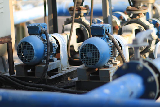 Several blue industrial electric motors are mounted on steel bases, connected to pipes and other machinery with electrical cables. The setting appears to be a mechanical or industrial facility with focused lighting on the motors and parts.