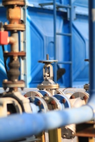 An industrial setup with numerous pipes and valves, primarily featuring shades of blue and metallic surfaces. The image captures a close-up of a valve in focus with a blurred background showing more industrial equipment and a blue structural element.