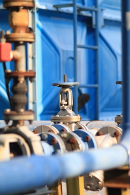 An industrial setup with numerous pipes and valves, primarily featuring shades of blue and metallic surfaces. The image captures a close-up of a valve in focus with a blurred background showing more industrial equipment and a blue structural element.