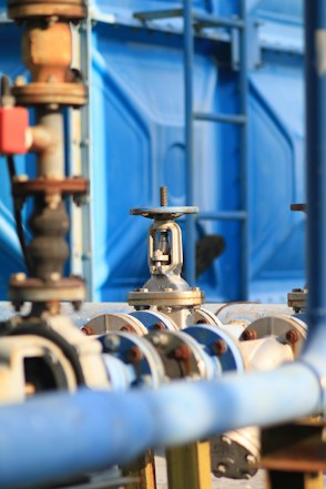 An industrial setup with numerous pipes and valves, primarily featuring shades of blue and metallic surfaces. The image captures a close-up of a valve in focus with a blurred background showing more industrial equipment and a blue structural element.