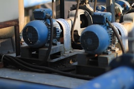 Two large blue industrial electric motors are mounted on metal frames. They are connected by black electrical cables, and the surrounding area contains various industrial components and piping. The motors appear to be part of a mechanical system used in a factory or plant setting.