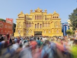 A large, ornate structure resembling a traditional palace is surrounded by a bustling crowd of people, indicating a festive or cultural event. The palace-like building features intricate architectural details and is predominantly golden in color. Banners and advertisements are visible in the foreground, with blurred movement suggesting a lively atmosphere.