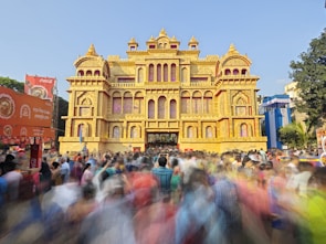 A large, ornate structure resembling a traditional palace is surrounded by a bustling crowd of people, indicating a festive or cultural event. The palace-like building features intricate architectural details and is predominantly golden in color. Banners and advertisements are visible in the foreground, with blurred movement suggesting a lively atmosphere.
