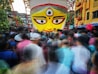A large, brightly colored depiction of a face with three eyes, a red bindi, and bold eyebrows is centered in the image. The area is crowded with people, creating a blur effect as they move around. The background features buildings and greenery.
