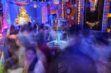 A vibrant and colorful festival scene filled with blurred images of people moving energetically around a decorated stage area. The stage features an ornate structure with vivid golden and orange colors, possibly a deity or symbolic figure. The surroundings are adorned with bright lights and intricate patterns, with clocks and circular designs on the walls, creating a lively, chaotic atmosphere.
