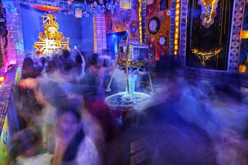 A vibrant and colorful festival scene filled with blurred images of people moving energetically around a decorated stage area. The stage features an ornate structure with vivid golden and orange colors, possibly a deity or symbolic figure. The surroundings are adorned with bright lights and intricate patterns, with clocks and circular designs on the walls, creating a lively, chaotic atmosphere.