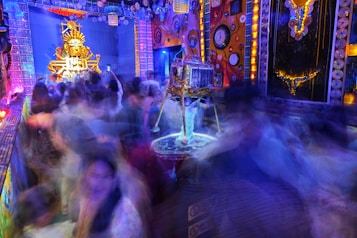 A vibrant and colorful festival scene filled with blurred images of people moving energetically around a decorated stage area. The stage features an ornate structure with vivid golden and orange colors, possibly a deity or symbolic figure. The surroundings are adorned with bright lights and intricate patterns, with clocks and circular designs on the walls, creating a lively, chaotic atmosphere.