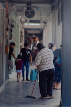 A person is sweeping the floor in a narrow corridor or hallway, while other people walk by in the background. The corridor is partially enclosed with visible light fixtures and shop fronts. There are a few individuals, including a child with an adult, moving along the passage.