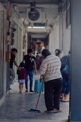 A person is sweeping the floor in a narrow corridor or hallway, while other people walk by in the background. The corridor is partially enclosed with visible light fixtures and shop fronts. There are a few individuals, including a child with an adult, moving along the passage.