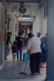 A person is sweeping the floor in a narrow corridor or hallway, while other people walk by in the background. The corridor is partially enclosed with visible light fixtures and shop fronts. There are a few individuals, including a child with an adult, moving along the passage.