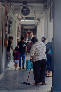 A person is sweeping the floor in a narrow corridor or hallway, while other people walk by in the background. The corridor is partially enclosed with visible light fixtures and shop fronts. There are a few individuals, including a child with an adult, moving along the passage.