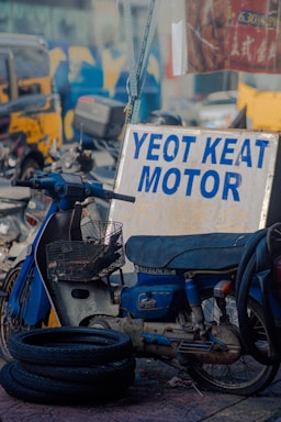 A vintage blue motorcycle parked next to a metal sign that reads 'Yeot Keat Motor'. The motorcycle appears worn, with a wire basket attached at the front. In the foreground, there are several worn rubber tires on the ground. The background features a blurred urban setting with various vehicles and colorful graffiti.