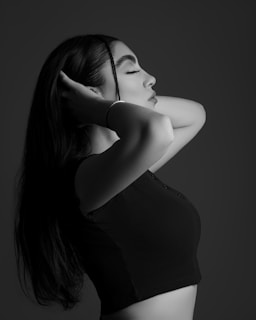 A black and white portrait of a person with long hair, eyes closed, and arms raised to touch their head, conveying a serene and contemplative mood.