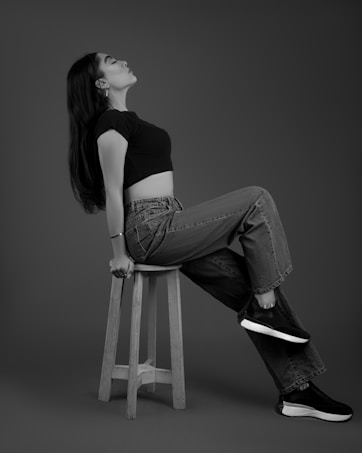 A black and white image of a person posing on a wooden stool. The individual is wearing a black top, jeans, and sneakers. They are leaning back slightly with their head tilted upwards, creating a relaxed and contemplative pose.