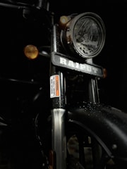 A close-up view of a motorcycle front showing a large headlamp with chrome details, a visible nameplate, and two amber turn signals. The metal surfaces are covered with water droplets, contributing to a wet appearance.