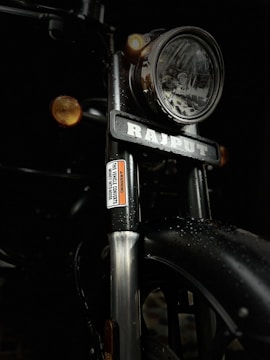 A close-up view of a motorcycle front showing a large headlamp with chrome details, a visible nameplate, and two amber turn signals. The metal surfaces are covered with water droplets, contributing to a wet appearance.