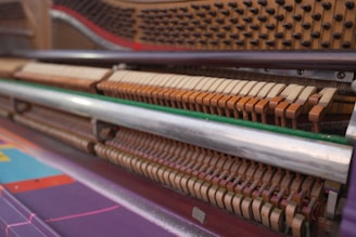 An up-close view of the internal mechanism of a piano, focusing on the hammers, strings, and dampers. The image shows the intricate details of the felt-topped hammers lined up in rows, ready to strike the strings when keys are pressed. The wooden components and metal strings are visible, with colorful elements visible at the bottom of the frame.