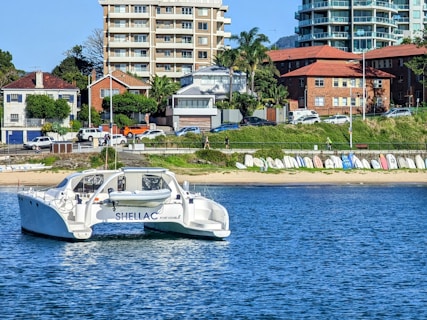 A catamaran with the name 'Shellac' is anchored near the shoreline, where a series of buildings including residential houses and taller apartment complexes are visible. The coastline features a sandy beach with a row of small rowboats or dinghies lined up on the shore. Trees and greenery are interspersed among the built structures, and a few cars are parked near the shore.