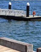 A floating dock extends into a body of water, secured by two black and white bollards. A lifebuoy is attached to one of the bollards for safety. The water is a deep blue with visible surface ripples, and the dock is bordered by a wooden railing.