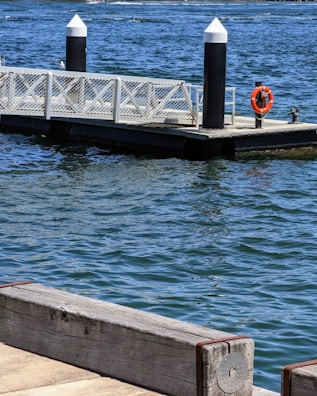 A floating dock extends into a body of water, secured by two black and white bollards. A lifebuoy is attached to one of the bollards for safety. The water is a deep blue with visible surface ripples, and the dock is bordered by a wooden railing.
