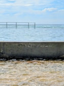 A tranquil ocean scene with a concrete barrier in the foreground, separating the calm waters from the rougher sea beyond. A narrow metal safety railing extends above the water, under a mostly clear sky with some clouds.