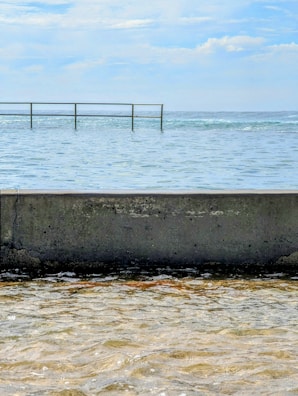 A tranquil ocean scene with a concrete barrier in the foreground, separating the calm waters from the rougher sea beyond. A narrow metal safety railing extends above the water, under a mostly clear sky with some clouds.